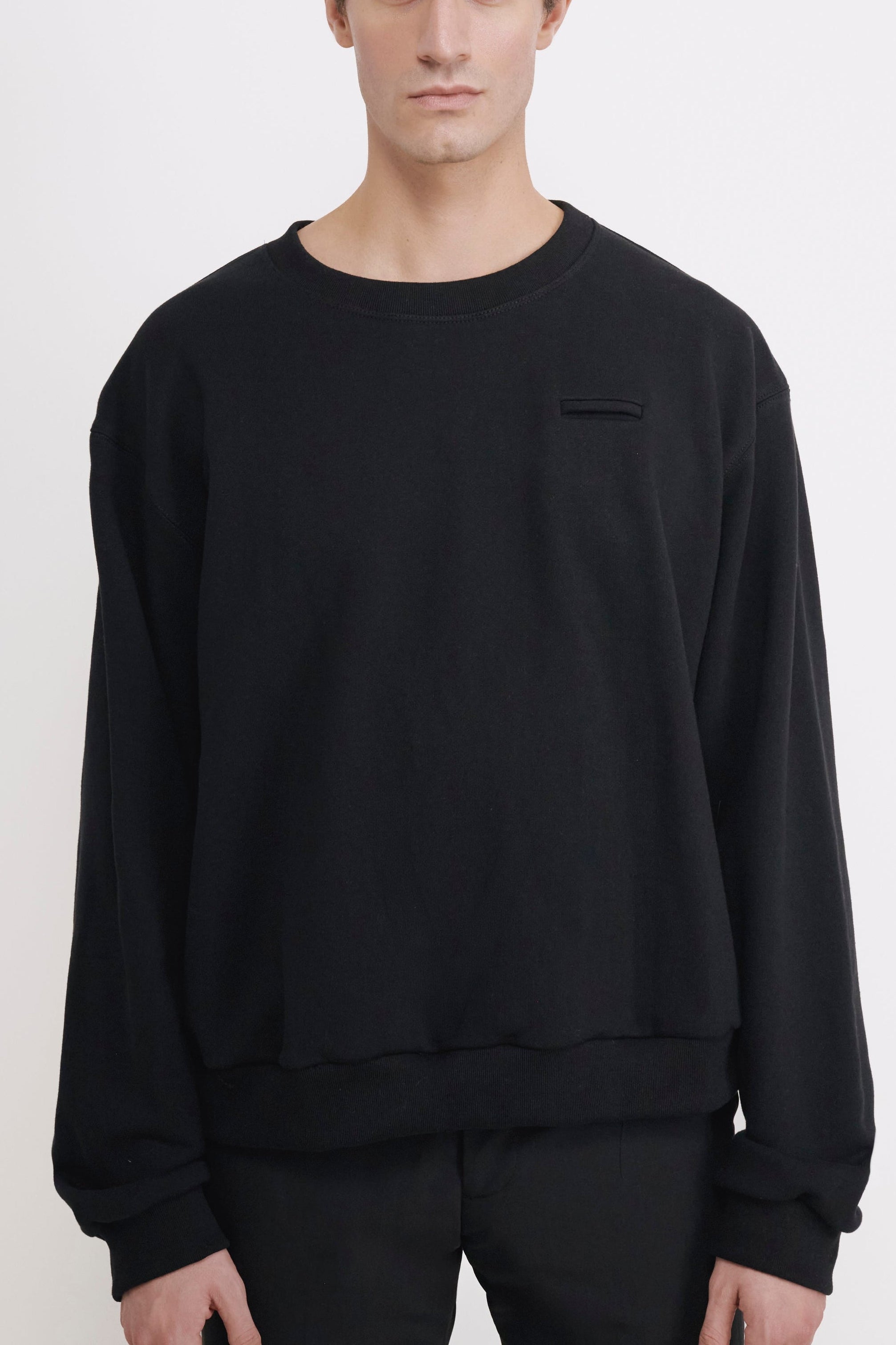 A man wearing a black crewneck sweatshirt with a small chest pocket and black pants, standing against a plain white studio background.
