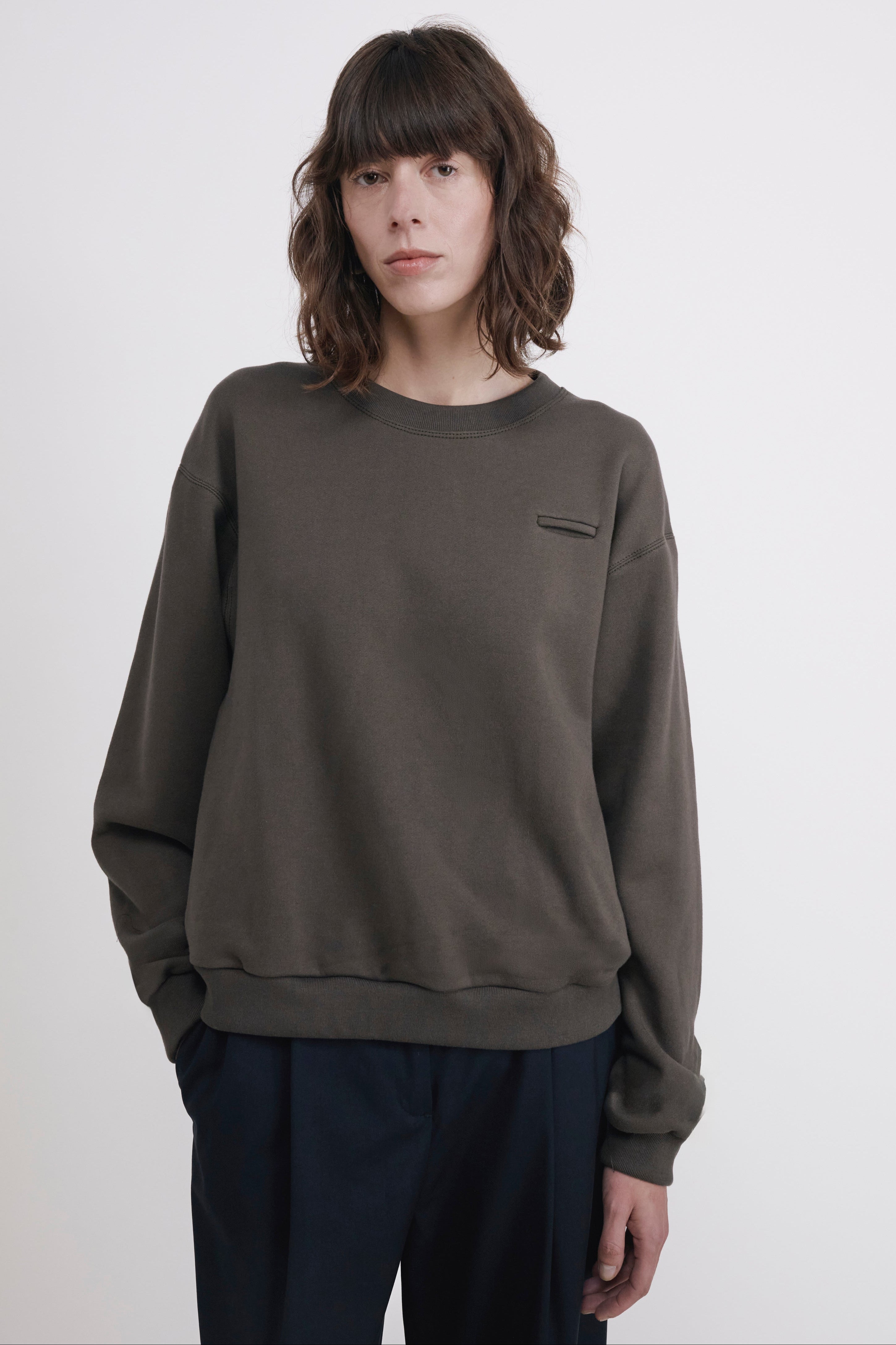 A model wearing a dark olive green crewneck sweatshirt with a small chest pocket and dark navy pants, standing against a plain light gray background.