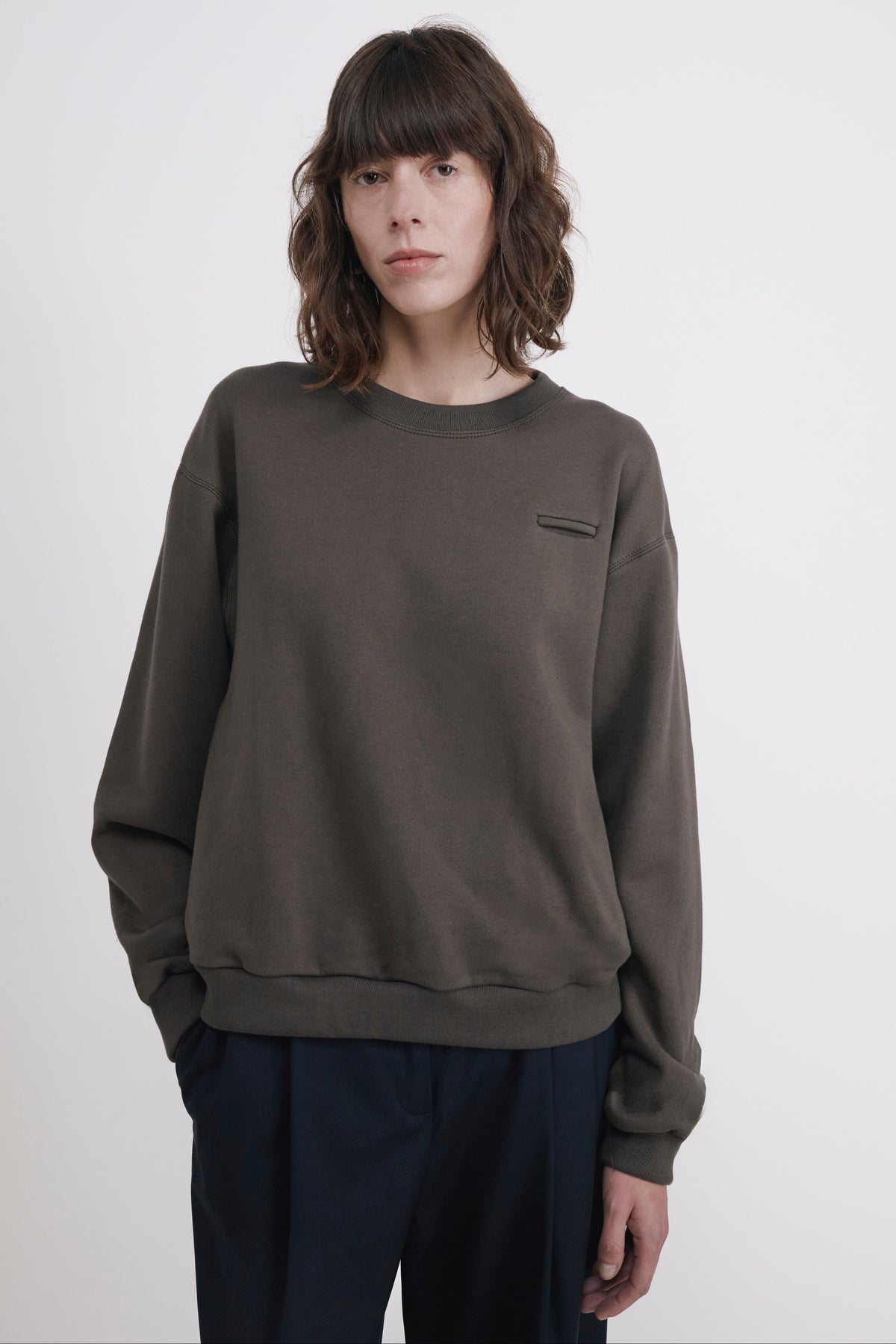 A model wearing a dark olive green crewneck sweatshirt with a small chest pocket and dark navy pants, standing against a plain light gray background.