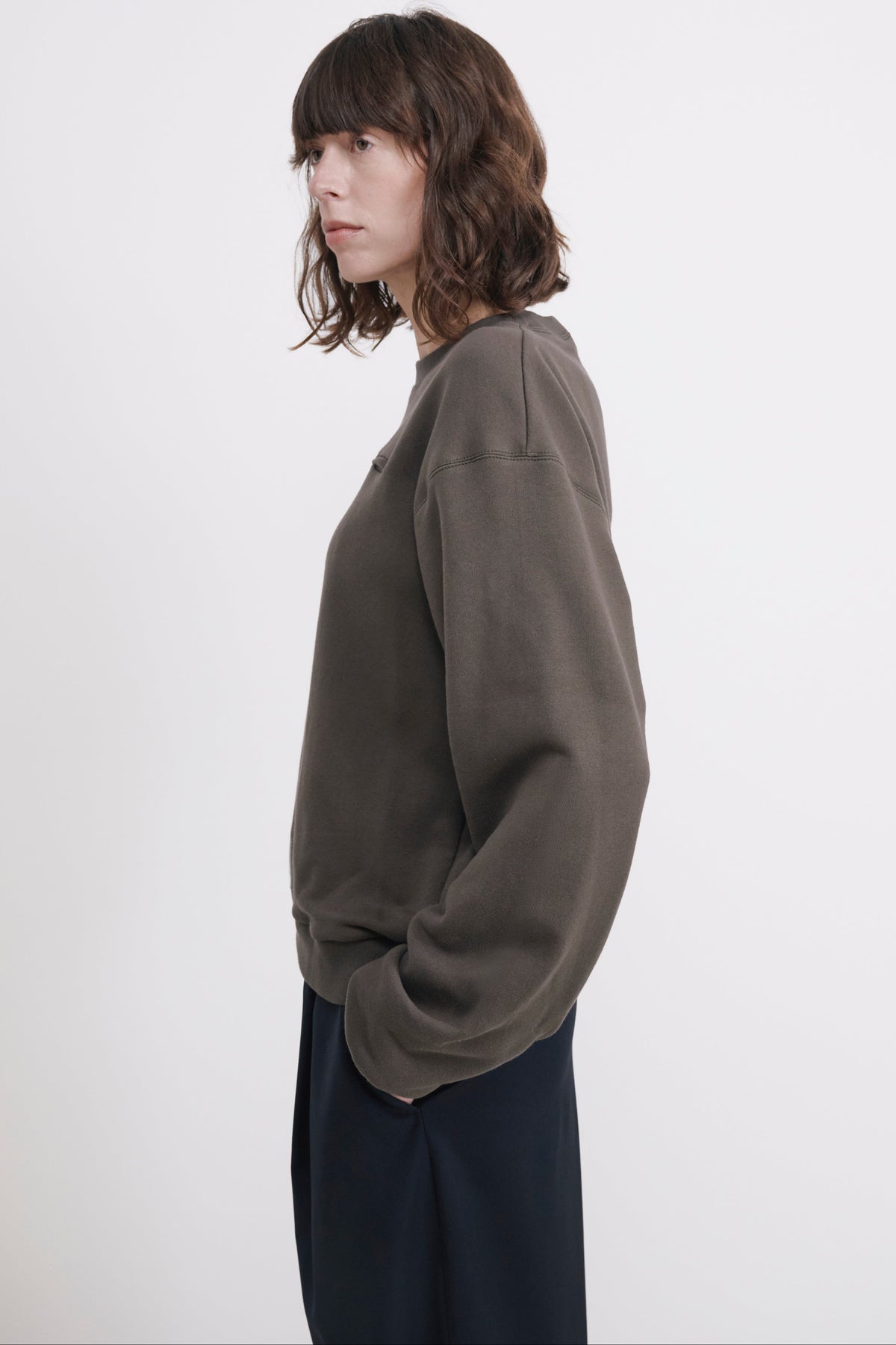A woman with shoulder-length brown hair is shown in profile wearing an oversized dark olive green sweatshirt and dark navy pants against a plain light gray studio background.