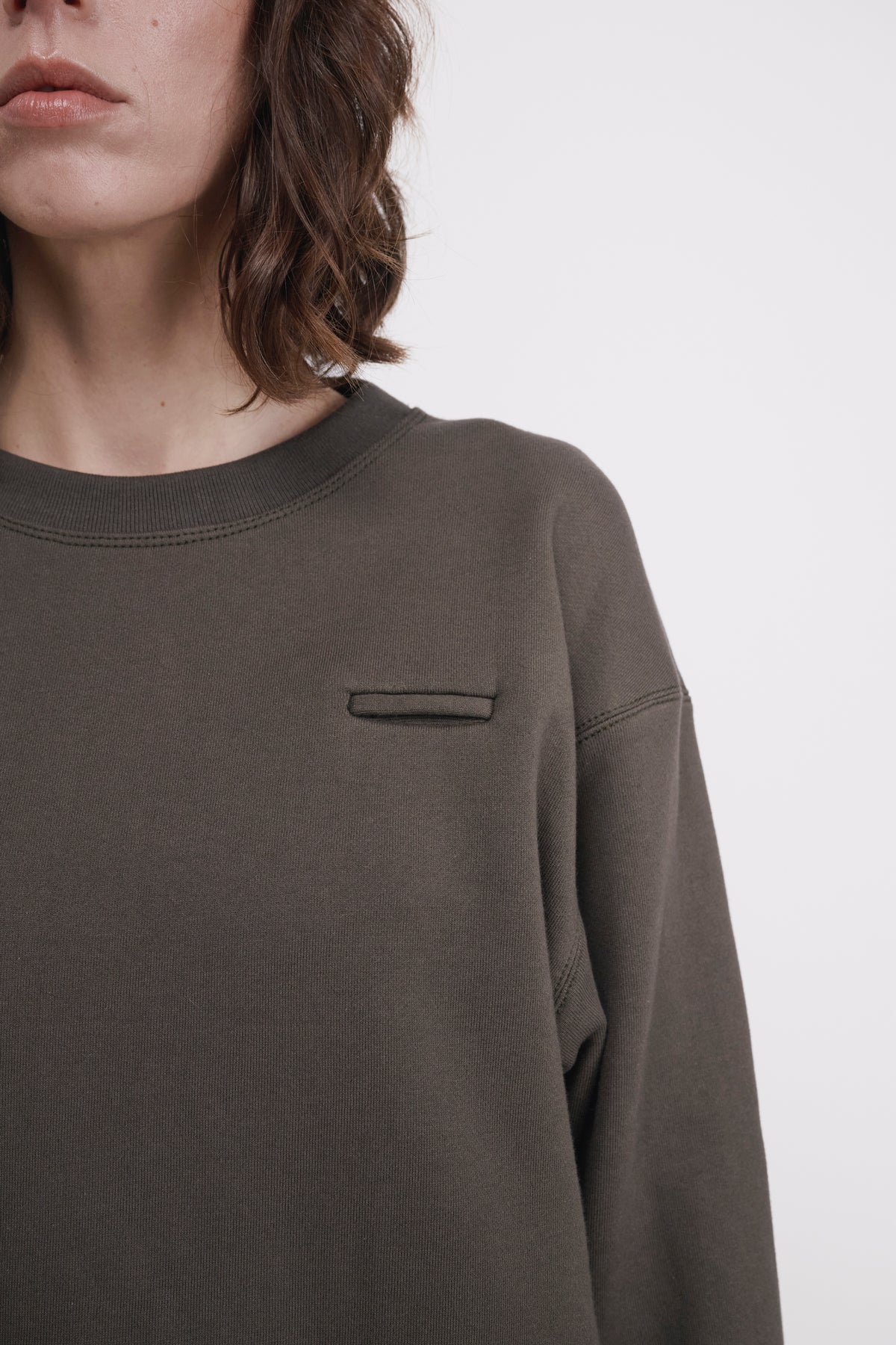 Close-up of a person wearing an oversized dark olive green crewneck sweatshirt with a small horizontal chest pocket, against a plain light gray background.
