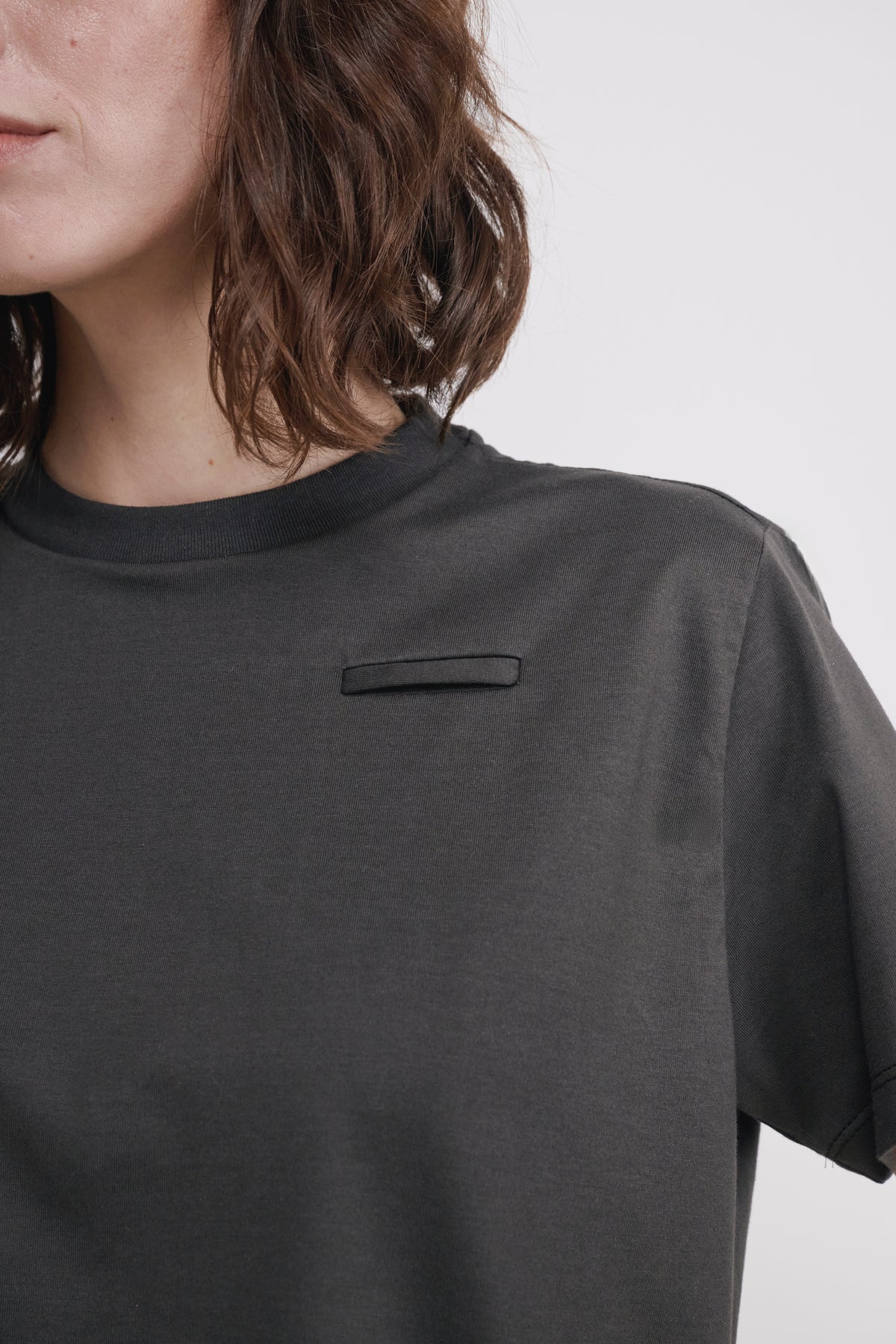 Close-up of a person wearing a dark charcoal gray short-sleeve t-shirt with a small rectangular patch detail on the chest, against a light gray background.