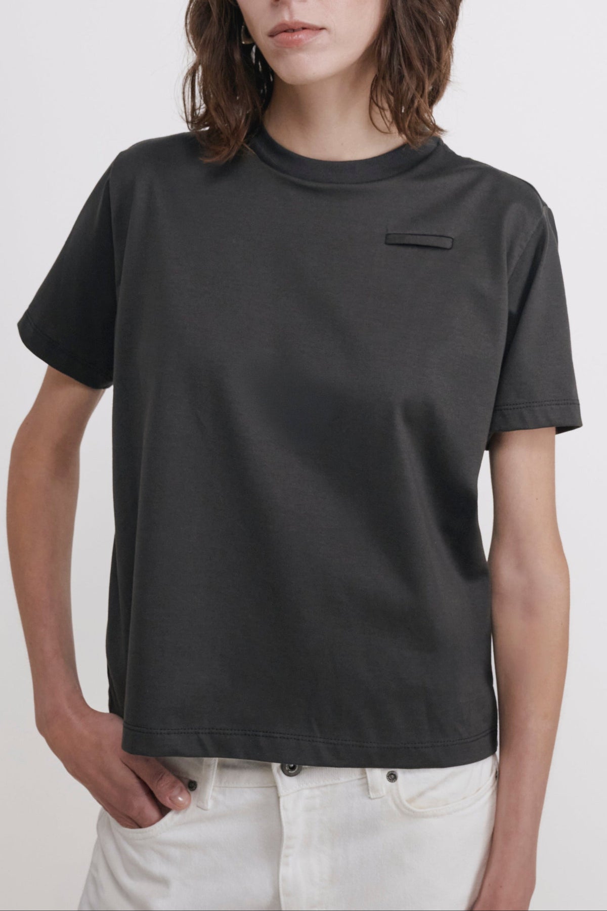 A model with shoulder-length brown hair wearing a dark charcoal gray short-sleeve t-shirt with a small chest pocket and white pants, standing against a plain light gray studio background.