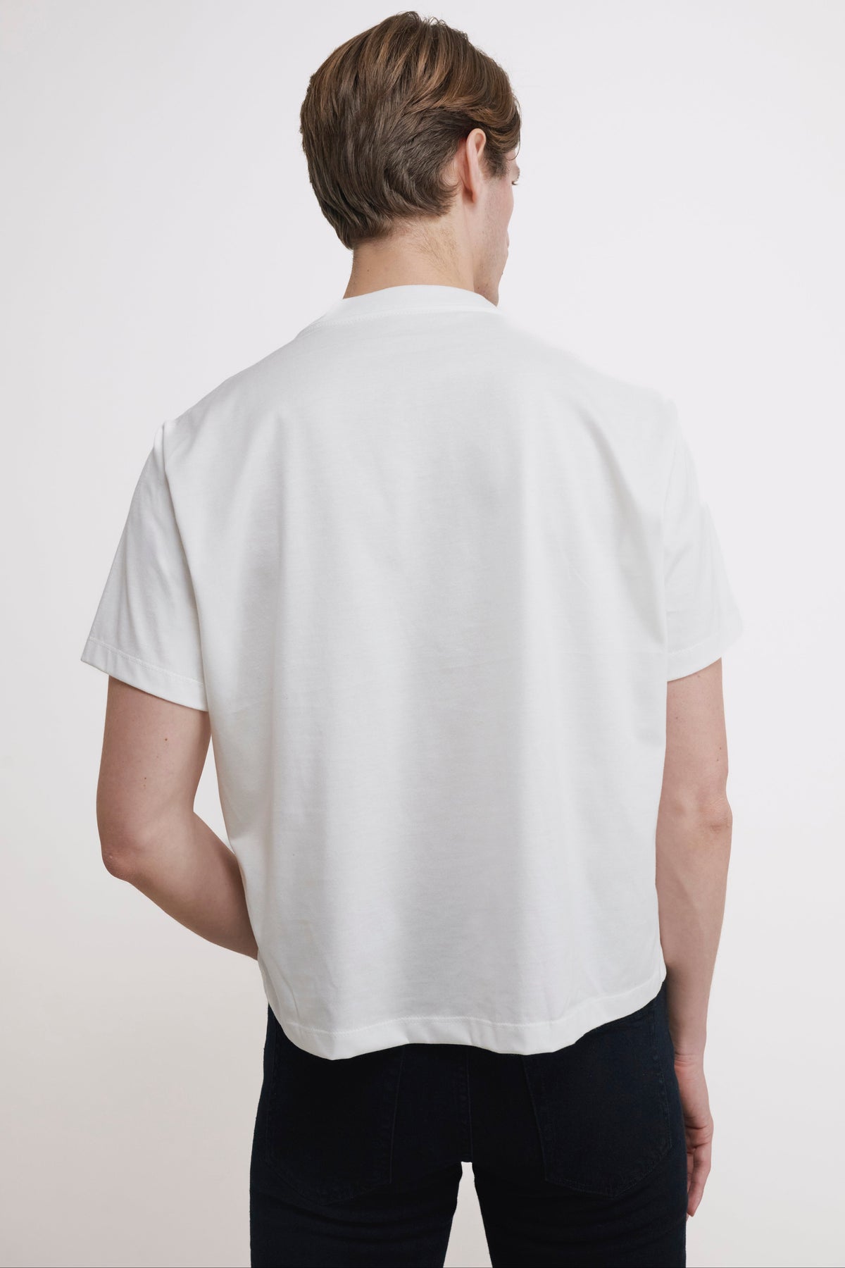 A man viewed from behind wearing a white short-sleeve t-shirt and black pants against a plain white studio background.