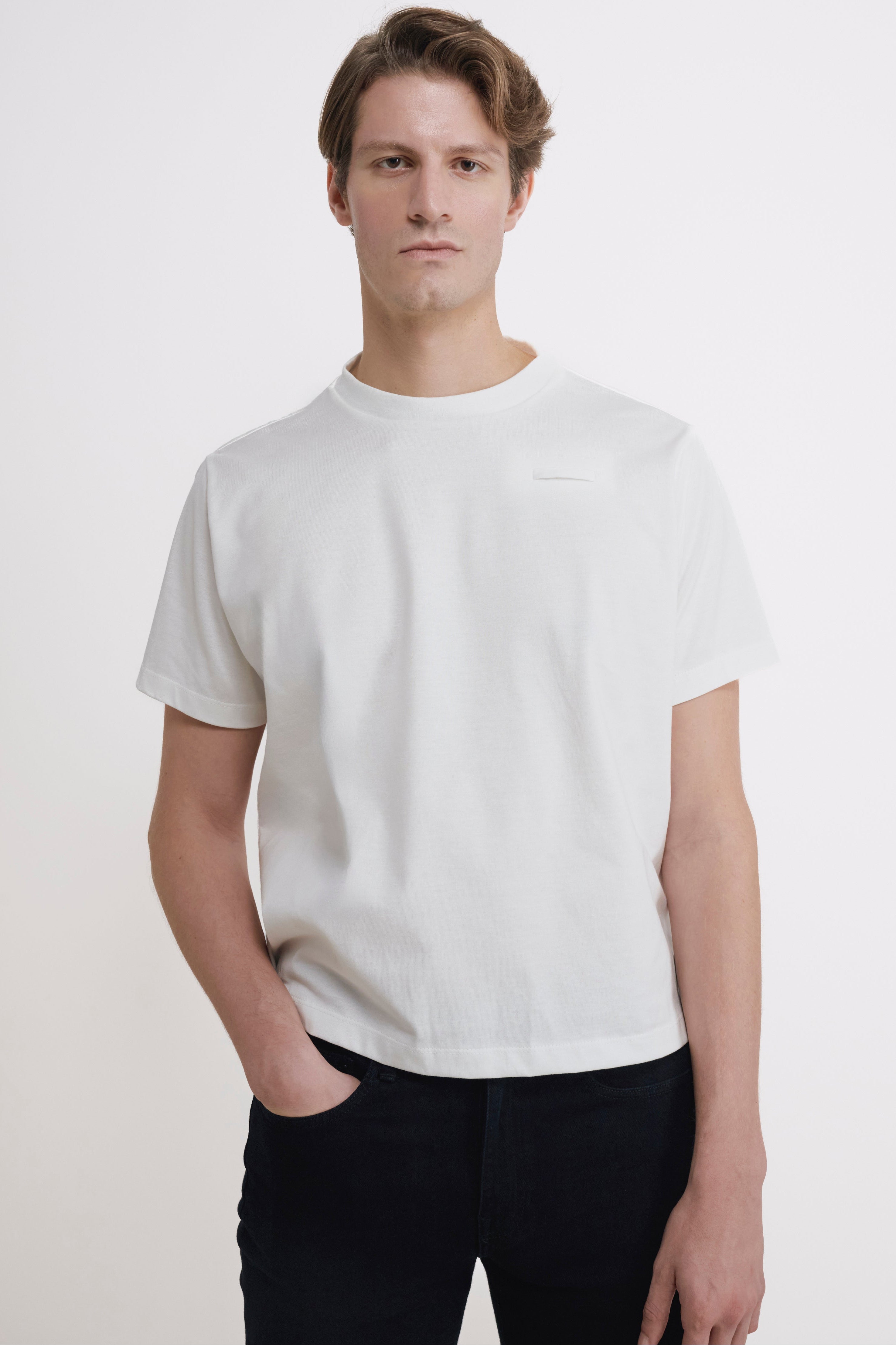 A man wearing a plain white short-sleeve crewneck t-shirt and black pants, standing against a light off-white studio background.