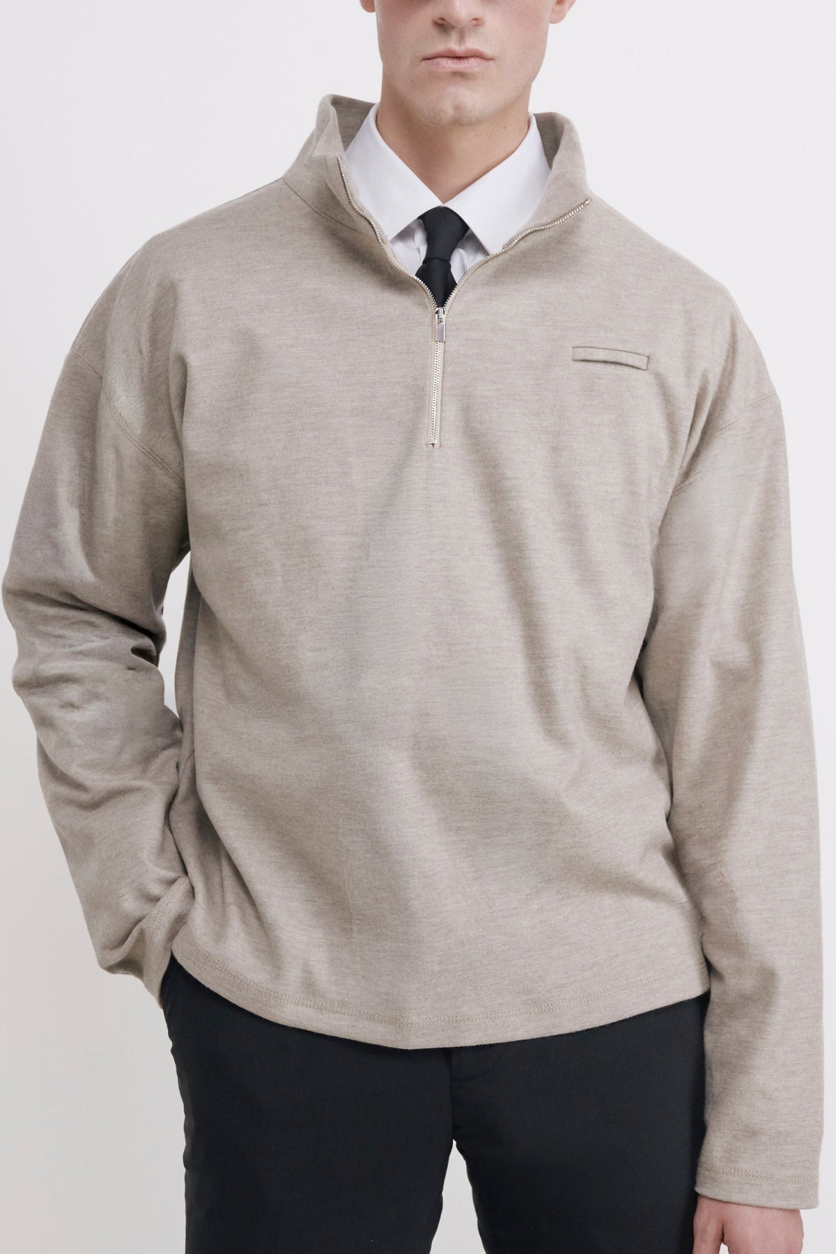 A man wearing a light beige quarter-zip pullover sweater over a white collared shirt and black tie, paired with black trousers, standing against a plain light gray studio background.