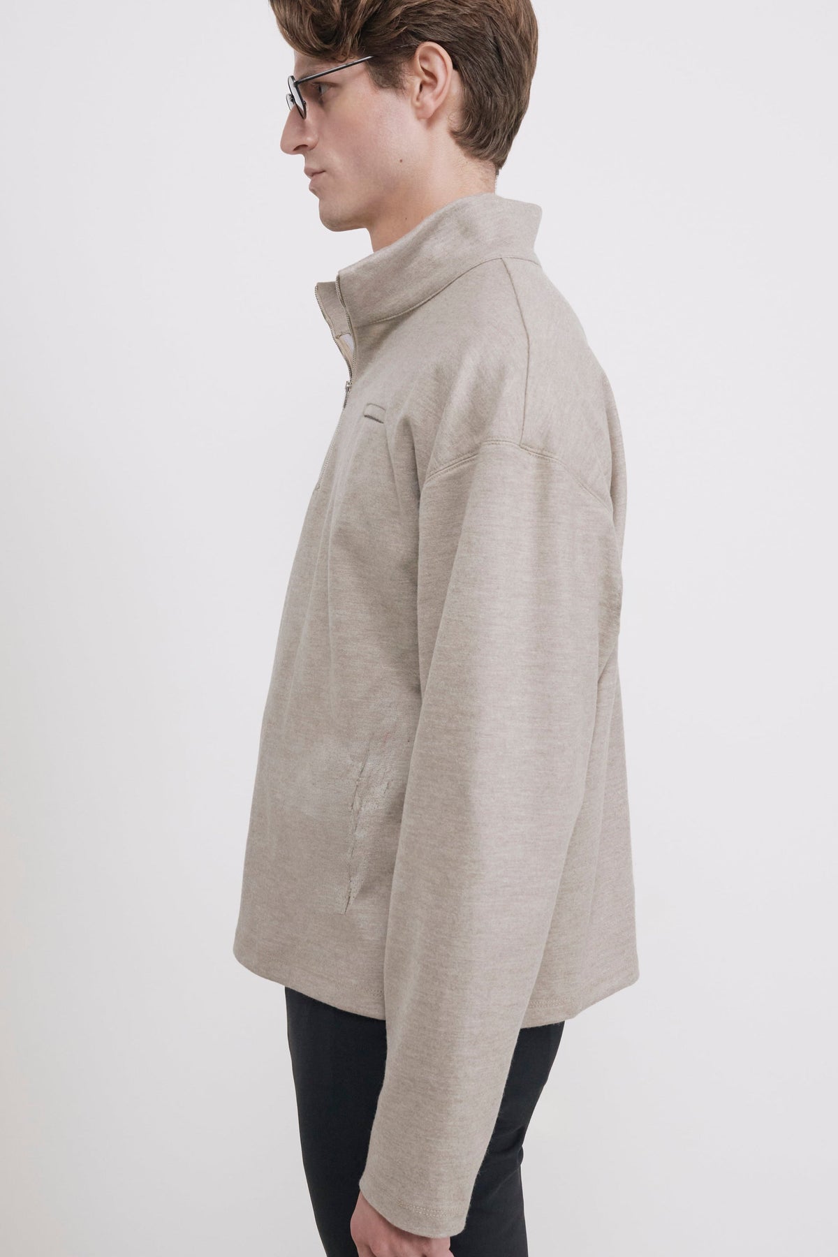 A man in profile wearing a light beige heathered quarter-zip pullover jacket and dark pants, standing against a plain light gray studio background.