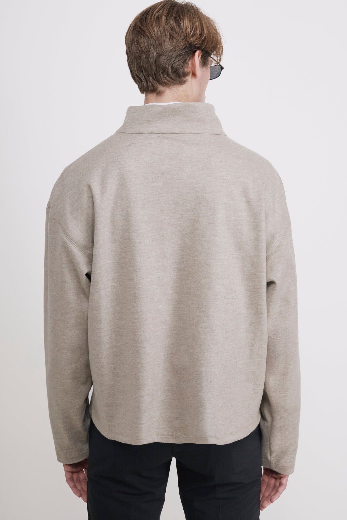 A man viewed from behind wearing a light beige mock-neck knit sweater and dark trousers, standing against a plain light gray studio background.