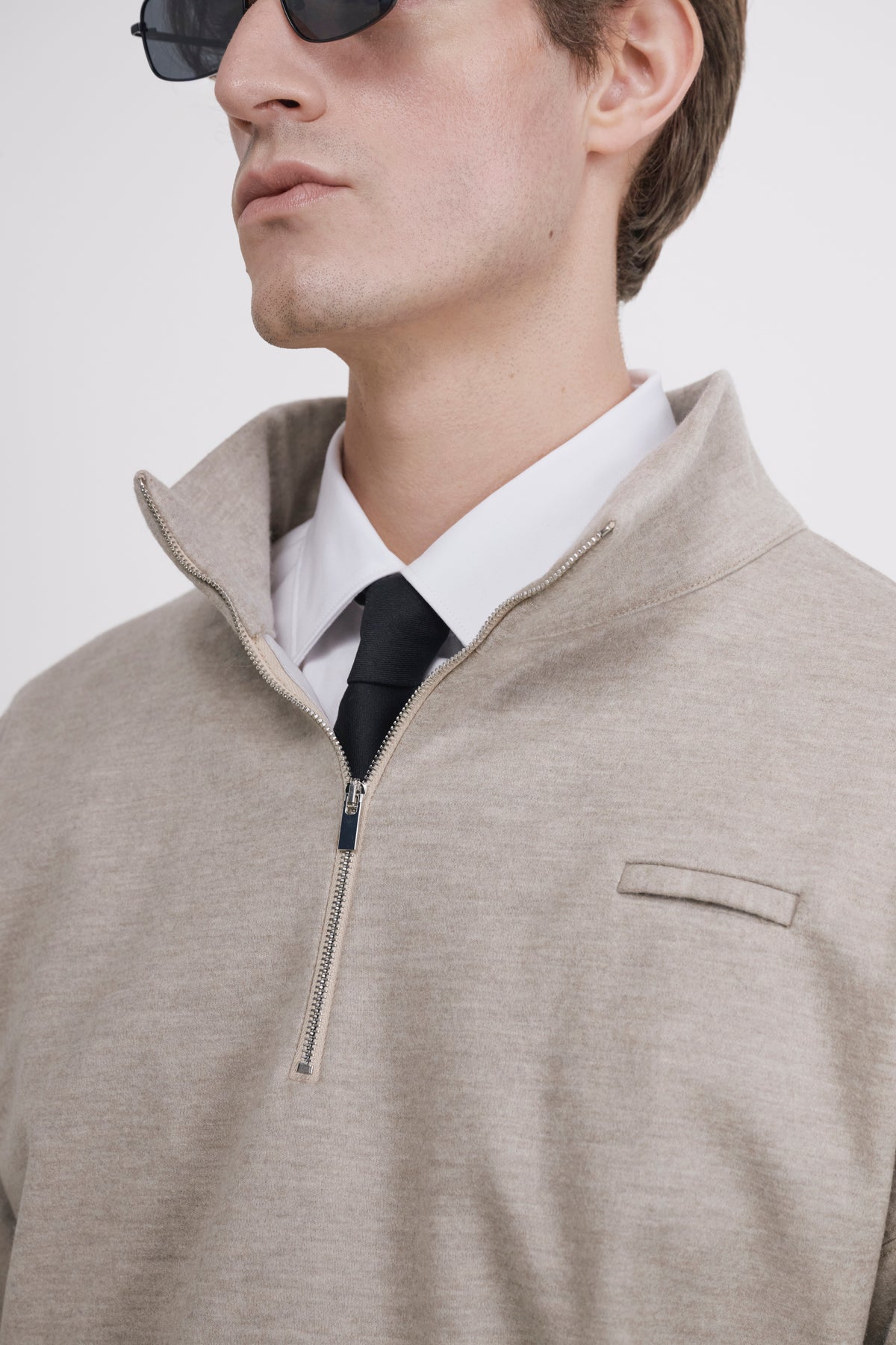 A close-up of a man wearing a light beige quarter-zip pullover sweater over a white collared shirt and black tie, with sunglasses partially visible.