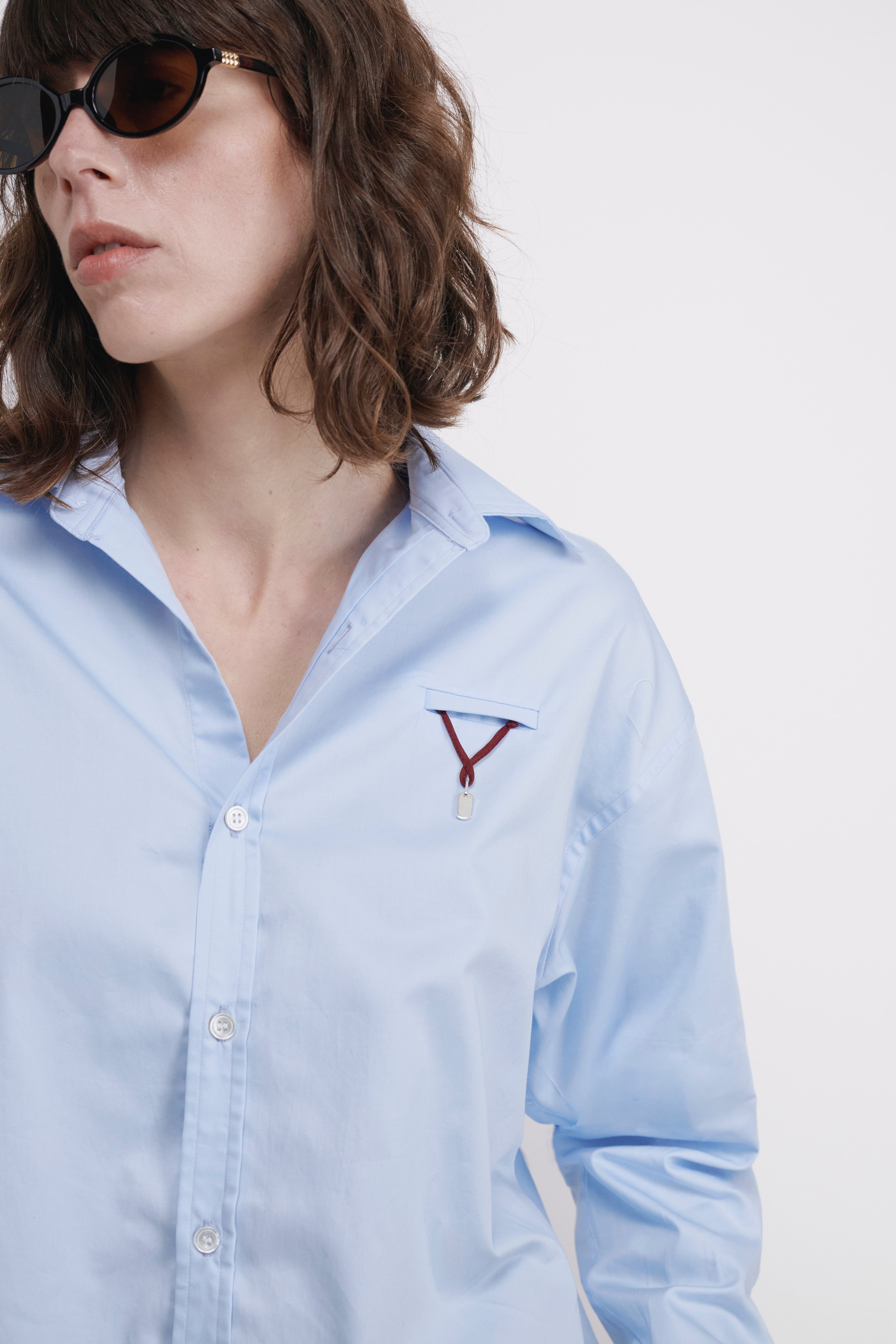 A close-up of a model wearing a light blue button-down shirt with a small embroidered red and white geometric logo on the chest, paired with dark sunglasses. Burgundy Attachment