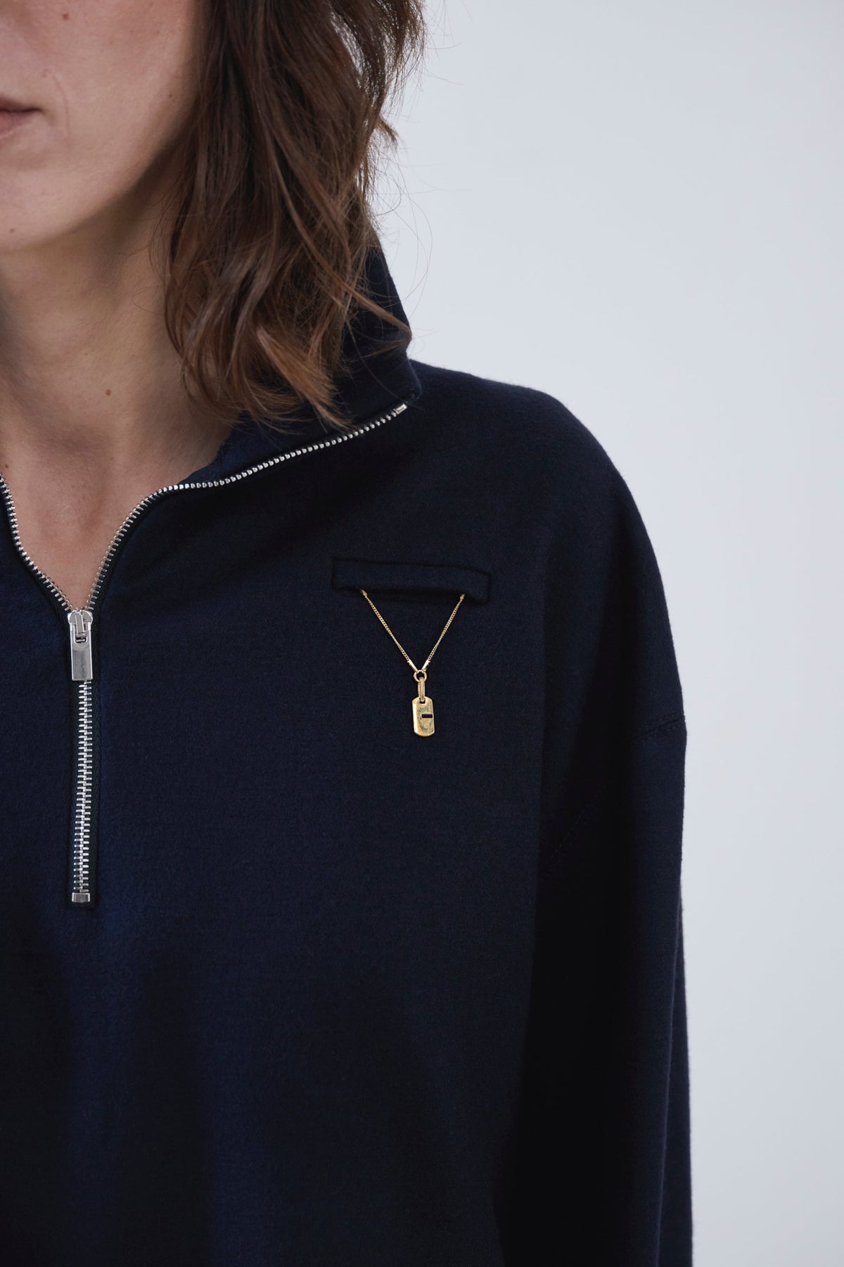 A close-up of a person wearing a dark navy quarter-zip pullover with a small gold pendant necklace pinned to the chest pocket.