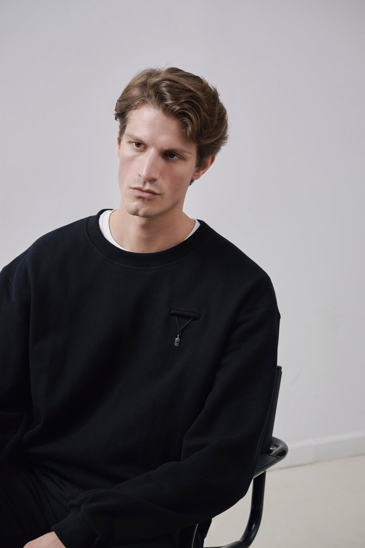 A man wearing a black crewneck sweatshirt with a small black Y-Logo on the chest, sitting on a black chair against a light gray studio background. Black Attachment