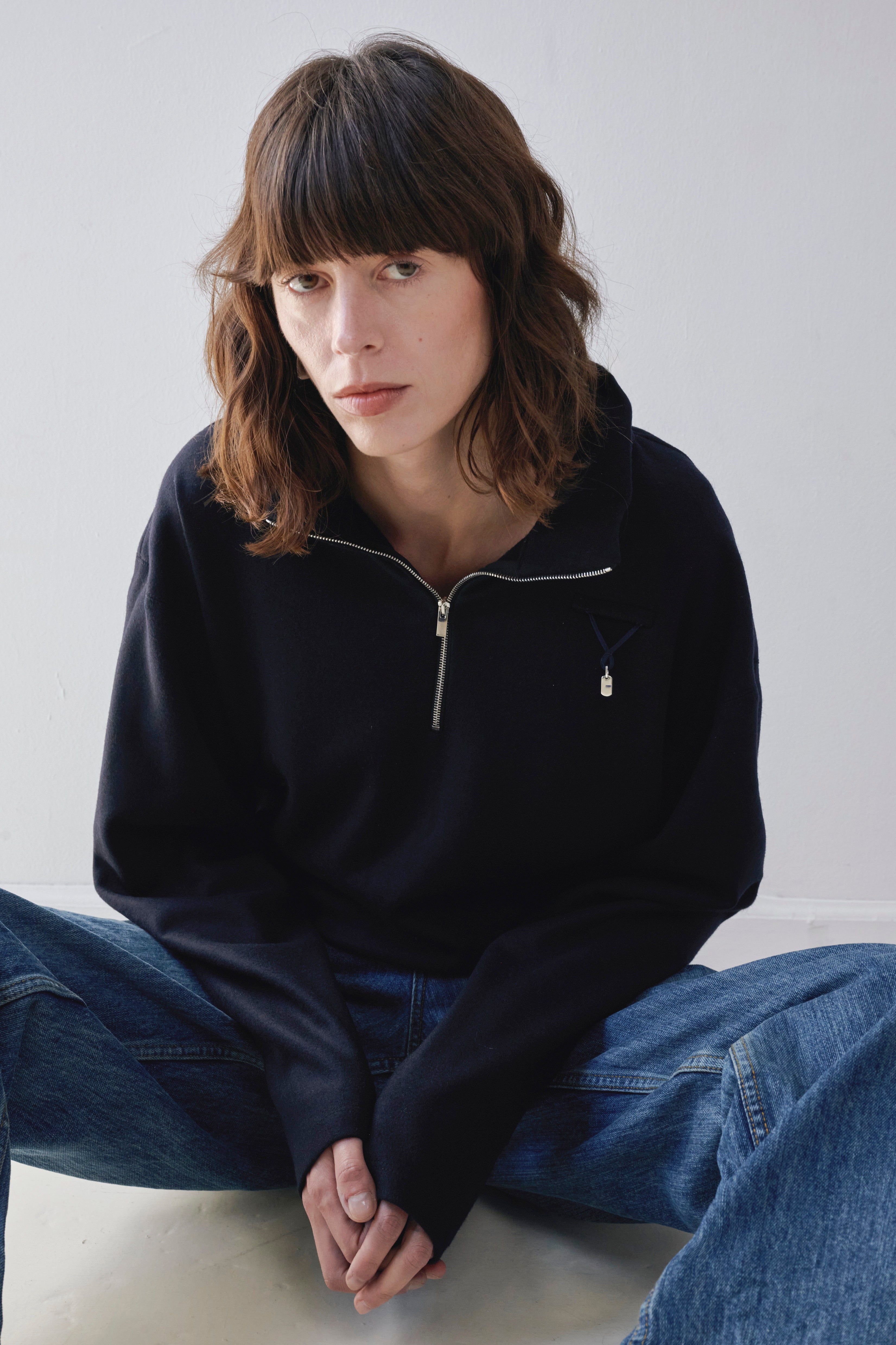 A woman with shoulder-length brown hair sits cross-legged against a plain light gray wall, wearing a navy half-zip pullover with a small silver zipper pull detail and blue jeans and navy attachment