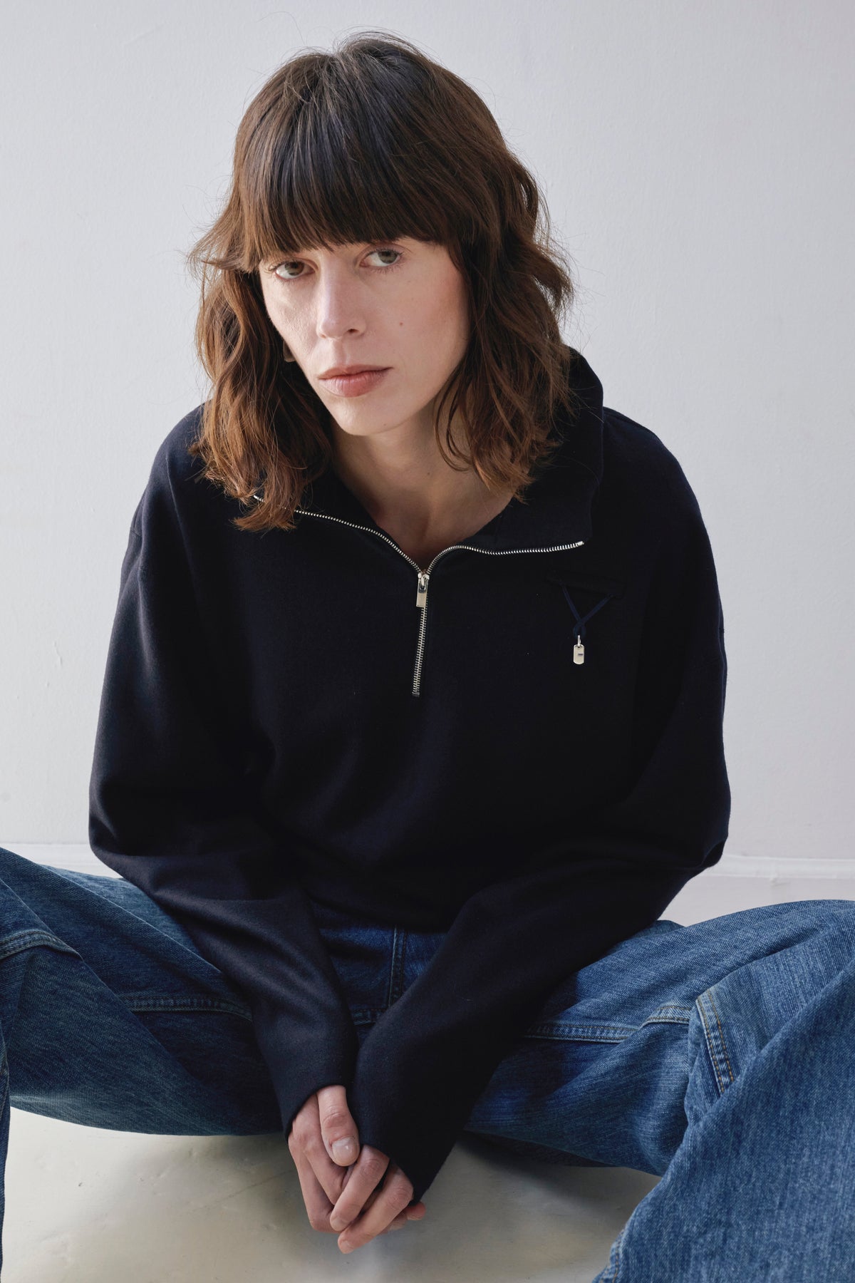 A woman with shoulder-length brown hair sits cross-legged against a plain light gray wall, wearing a navy half-zip pullover with a small silver zipper pull detail and blue jeans and navy attachment